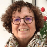 Portrait souriant d'une femme aux cheveux bouclés châtains et aux lunettes rondes, devant un mur clair.