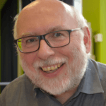 Portrait souriant d'un homme à la barbe blanche et aux lunettes.