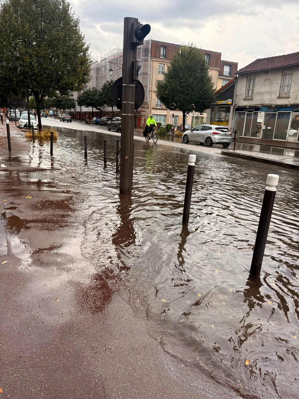 Une chaussée et un trottoir recouverts de plusieurs centimètres d’eau autour de bornes et d’un mât de feu tricolore. Au fond, une personne à vélo en veste jaune fluo traverse l’eau, devant des commerces et un immeuble sous échafaudage.