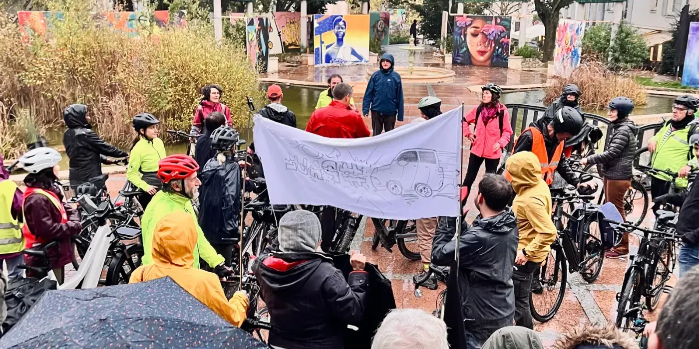 Une centaine de personnes rassemblées sous la pluie, beaucoup à vélo et en casque, autour d'une banderole blanche tendue sur laquelle un SUV est dessiné.