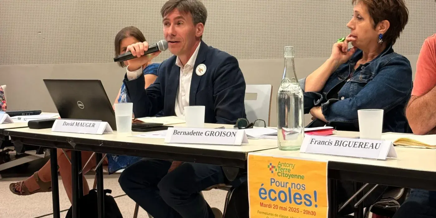 Table des intervenants de la soirée-débat, un homme au micro, les cartons nominatifs de David Mauger, Bernadette Groison et Francis Biguereau posés devant eux, et l'affiche « Pour nos écoles ! » accrochée au plateau.